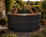 Two people relaxing in a large round outdoor stock tank in a garden setting at sunset.