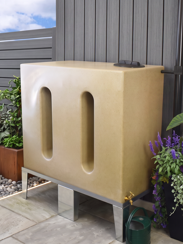 A modern outdoor rain barrel in a warm sandstone finish, on a metal stand in a landscaped backyard garden setting with plants and a gray fence.