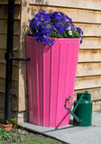 Mini Cosmo Water Butt Planter in Pink with purple flowers on a paved area.