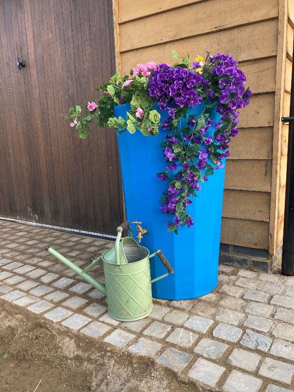 bright sky blue water butt planter on a stone patio, overflowing with purple and pink  flowers.