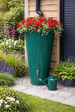 Green water butt planter with red flowers and a watering can in a garden setting.