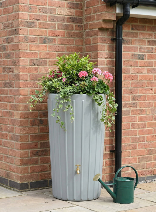Gray rain barrel with plants and a green watering can against a brick wall.
