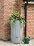 Gray rain barrel with plants and a green watering can against a brick wall.