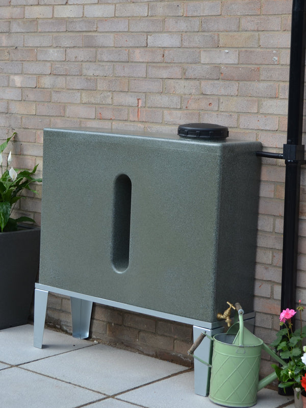 Green rain barrel attached to a brick wall with plants and a watering can in the foreground.