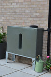 Green rain barrel attached to a brick wall with plants and a watering can in the foreground.