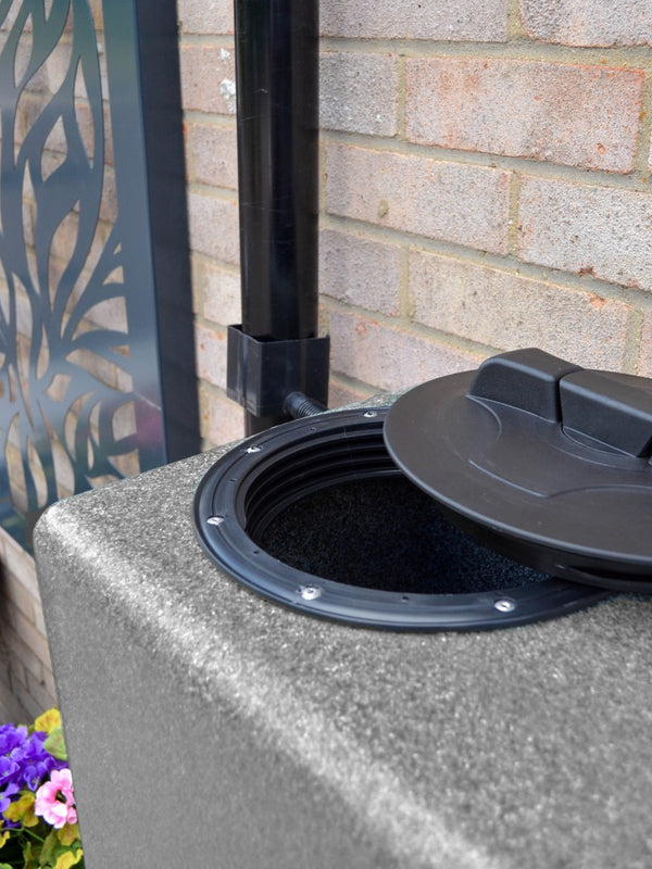 Rainwater harvesting system with a black overflow pipe and lid on a concrete block against a brick wall.