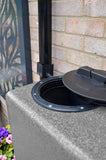 Rainwater harvesting system with a black overflow pipe and lid on a concrete block against a brick wall.